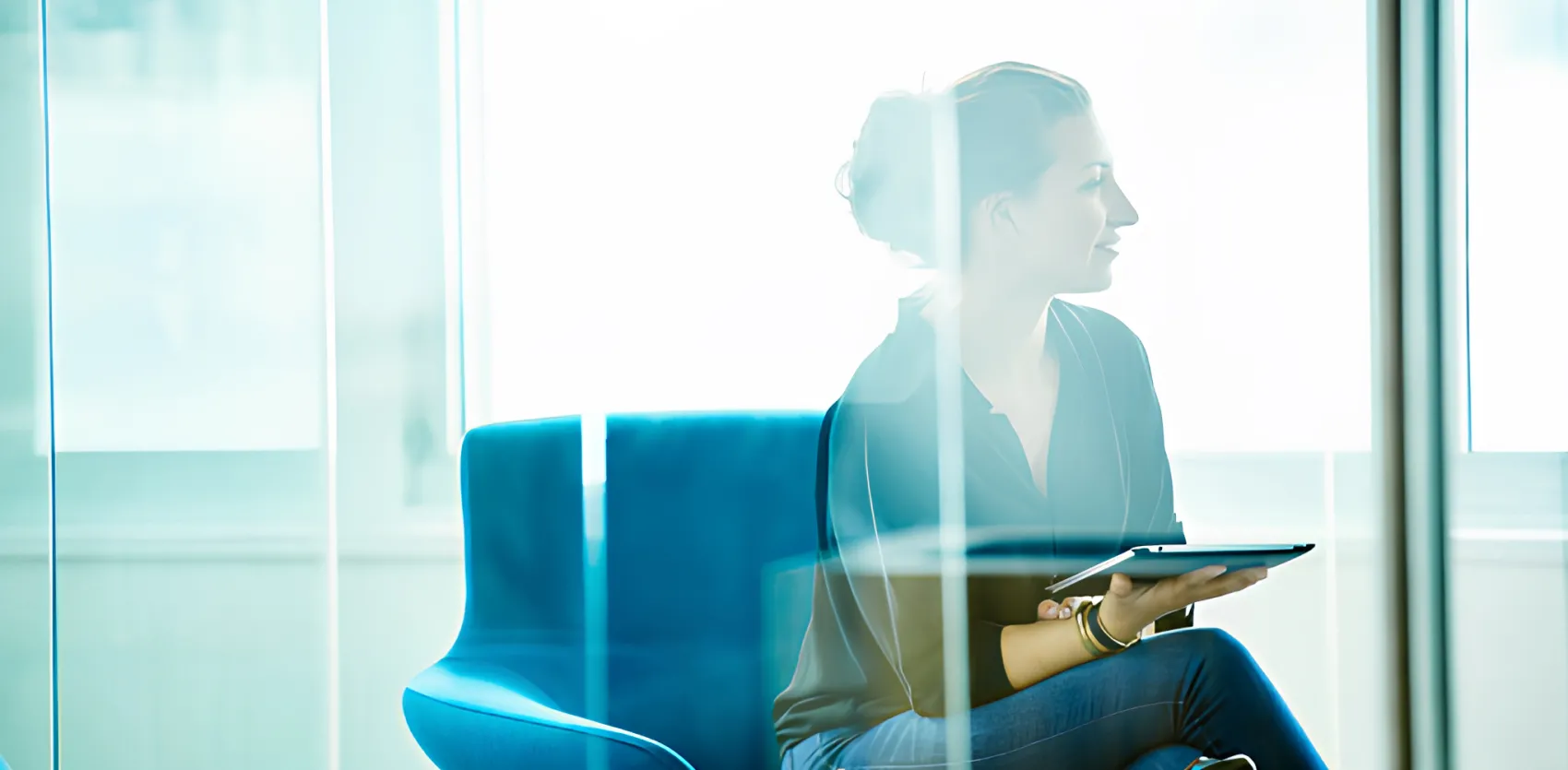 A woman sitting on a blue armchair, holding a tablet device, in an office environment