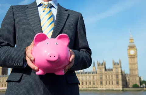 A man in a suit holding a piggy bank in front of the houses of parliament