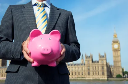 A man in a suit holding a piggy bank in front of the houses of parliament